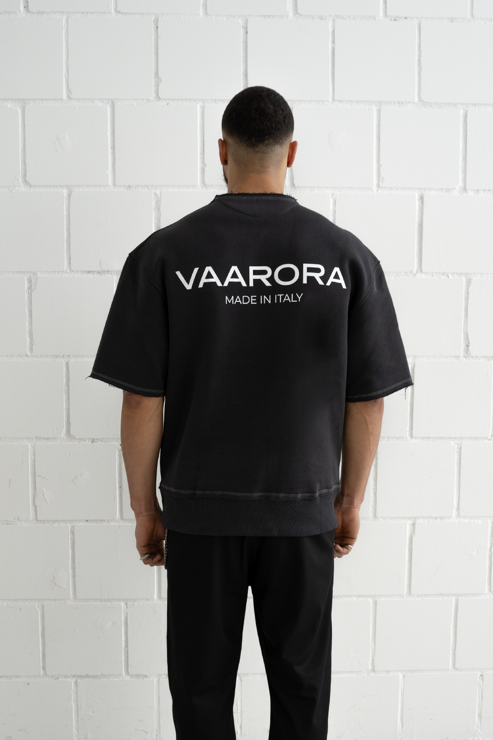 Back view of a male model wearing the black Vaarora Cato Heavy Tee featuring an oversized fit and bold white Made in Italy logo print